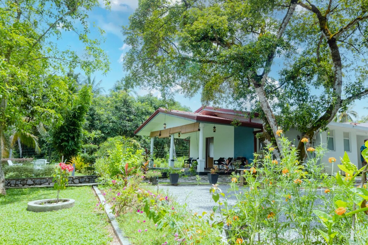 The exterior of the retreat is surrounded by vibrant gardens featuring a variety of plants and flowers. A sheltered seating area with tables and chairs is visible, positioned under a canopy of lush trees that provide dappled sunlight and a serene atmosphere.