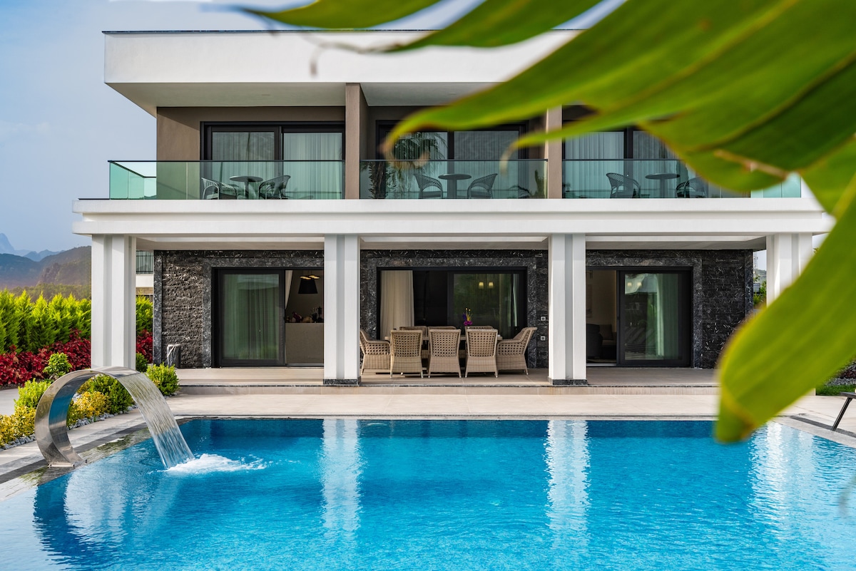 A modern villa is showcased with a private pool surrounded by landscaped gardens. A gentle water feature adds a tranquil element. Large glass doors open to the living area, enhancing the connection between indoor and outdoor spaces. Comfortable seating is visible on the patio.