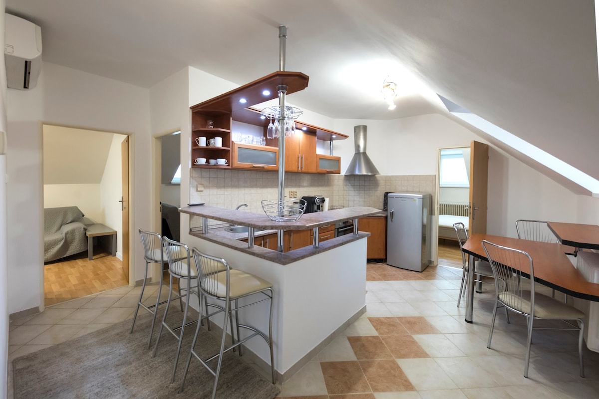 The open-concept kitchen features wooden cabinets and modern appliances, including a refrigerator and stovetop. A spacious dining area with a wooden table and metal chairs provides seating. Soft lighting enhances the relaxed atmosphere. A cozy seating area is visible through the doorway.