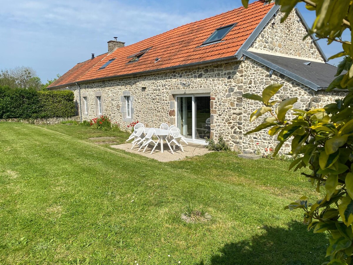 A charming stone house is presented with a red-tiled roof, set in a spacious grassy area. A patio is visible, furnished with white lounge chairs, providing a relaxed outdoor setting. Surrounding greenery enhances the peaceful ambiance of the property.