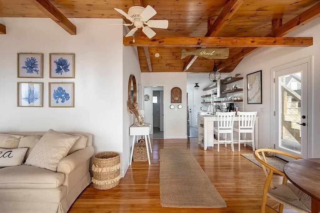 A light-filled living space features a comfortable sofa accompanied by decorative pillows. Natural wood beams and a ceiling fan enhance the airy atmosphere. A rug leads to a dining area with high chairs and an open-concept kitchen visible in the background.