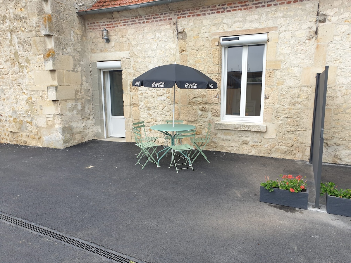 An outdoor terrace area is featured, showcasing a round table with four green chairs under an umbrella. The surrounding stone wall adds character, while a planter with blooming flowers contributes a touch of color to the setting. Access to a door leads inside.