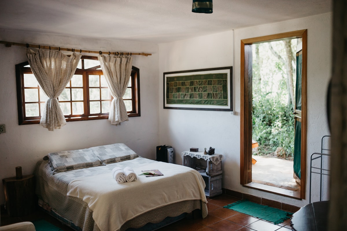 A bright and spacious sleeping area features a large bed with fresh linens. Soft natural light enters through two wooden windows, adorned with light curtains. A simple seating area is placed near the entrance, leading to a view of the surrounding greenery.