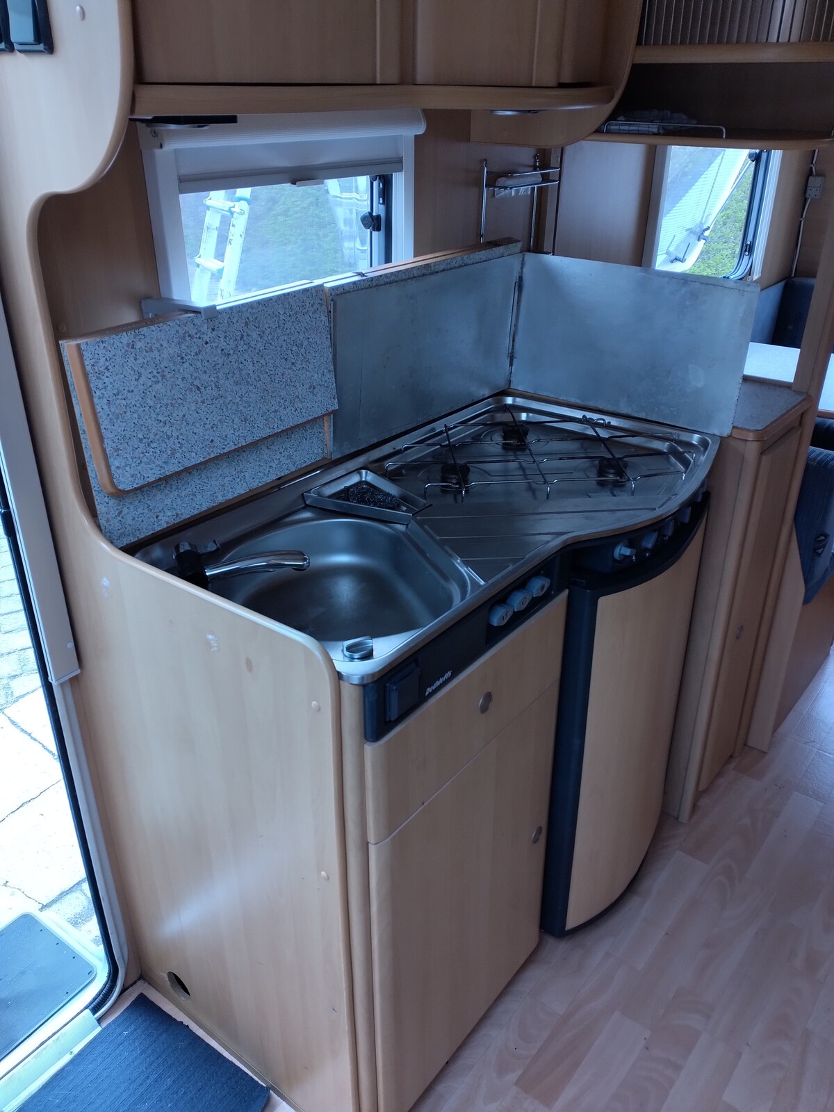 A compact kitchenette is visible, featuring a double burner gas stove and a stainless steel sink. Cabinets offer storage space, while a window provides natural light. The countertops are finished in patterned grey, complementing the wooden cabinetry.