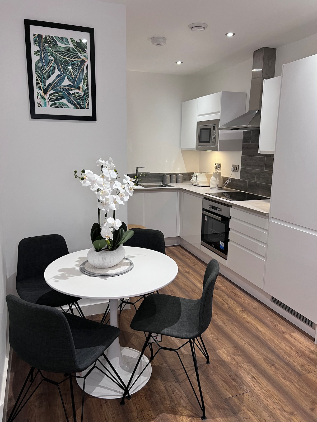 A modern kitchen area is visible, featuring sleek white cabinetry and stainless steel appliances. A round white table is set for four, accompanied by black chairs. A decorative plant arrangement adds a touch of elegance, while a wall-mounted artwork complements the space.