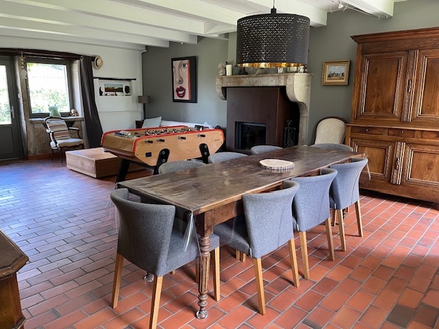 A spacious living area features a large wooden dining table surrounded by six upholstered chairs, with a brightly colored foosball table nearby. A stone fireplace and a vintage wooden cabinet are also present, against a backdrop of warm-toned tiled flooring.