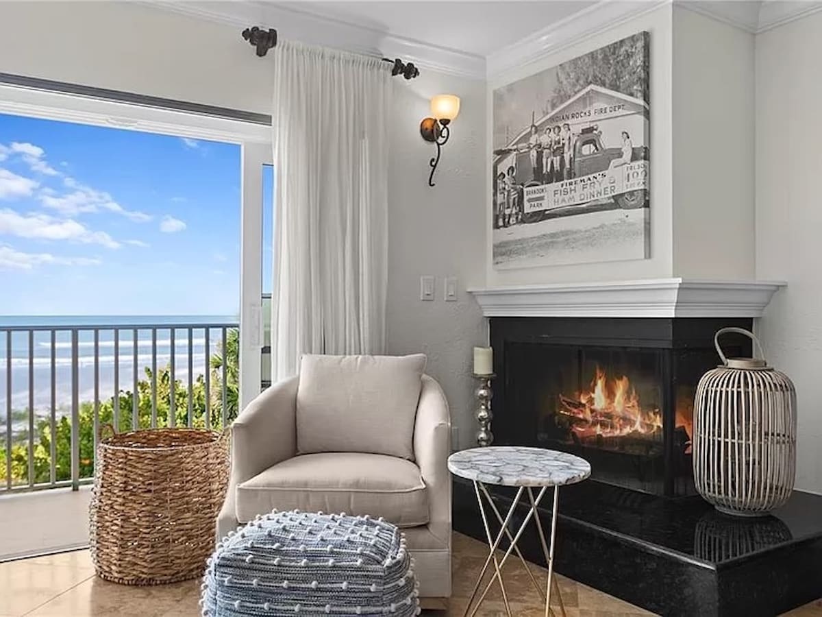 A cozy seating area features a plush armchair positioned near a gas fireplace, with a small table alongside. Large windows offer a view of the Gulf, while decorative elements include a woven basket and a textured pouf, adding warmth to the space.