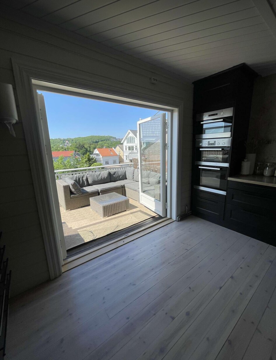 A doorway opens to a spacious terrace featuring outdoor seating, with views of the surrounding landscape. Sunlight floods the interior, highlighting the light wood flooring and modern kitchen appliances that are integrated within the sleek cabinetry.