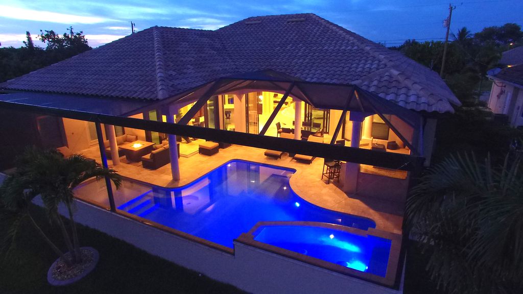 An aerial view shows the home's covered outdoor area featuring a spacious pool illuminated in blue. Surrounding the pool are lounge chairs and palm trees, creating a tranquil setting. The interior can be seen through large glass doors, emphasizing the open layout.