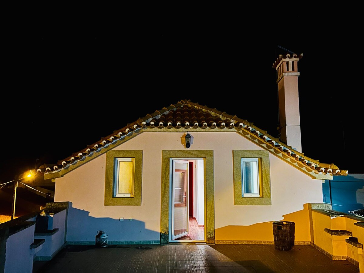 A charming building is showcased under a night sky, featuring a distinctive tiled roof and a welcoming entrance. Two windows are visible, allowing soft light to spill out, while a chimney adds character to the exterior.