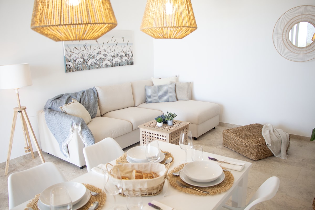 A modern living space is highlighted by a spacious sectional sofa adorned with soft pillows. Natural light illuminates the area, which features a dining table set for four and woven light fixtures above. A decorative plant adds a touch of greenery to the room.