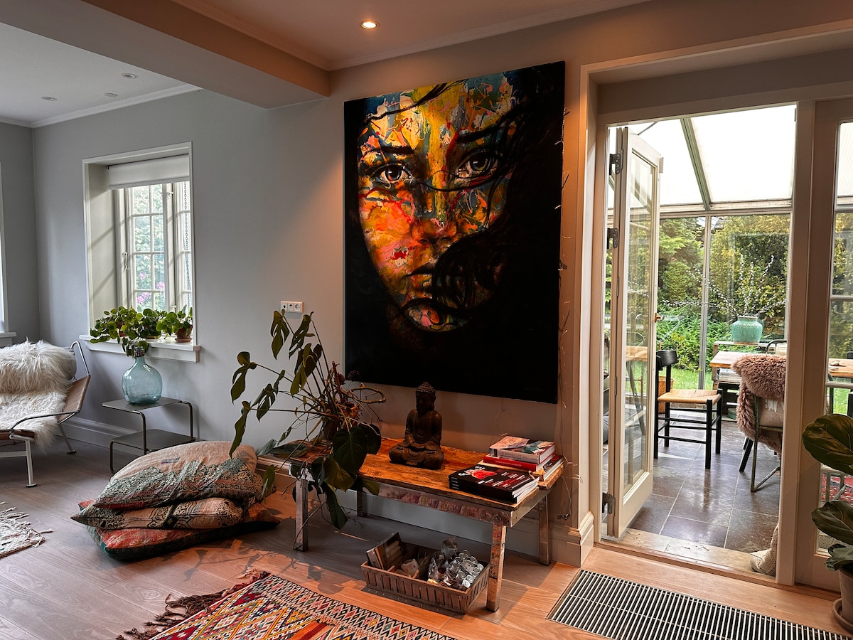 A vibrant painting of a woman's face serves as a focal point on the wall, showcasing colorful details. Natural light filters through glass doors leading to a garden space. Soft seating and decorative elements are arranged in a cozy living area, enhancing the inviting atmosphere.