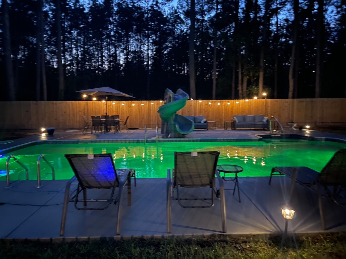 A large pool glows in green, surrounded by a spacious deck featuring lounge chairs. A water slide is positioned at the shallow end, and an outdoor dining area is visible in the background. Soft lighting enhances the ambiance against a backdrop of trees at dusk.