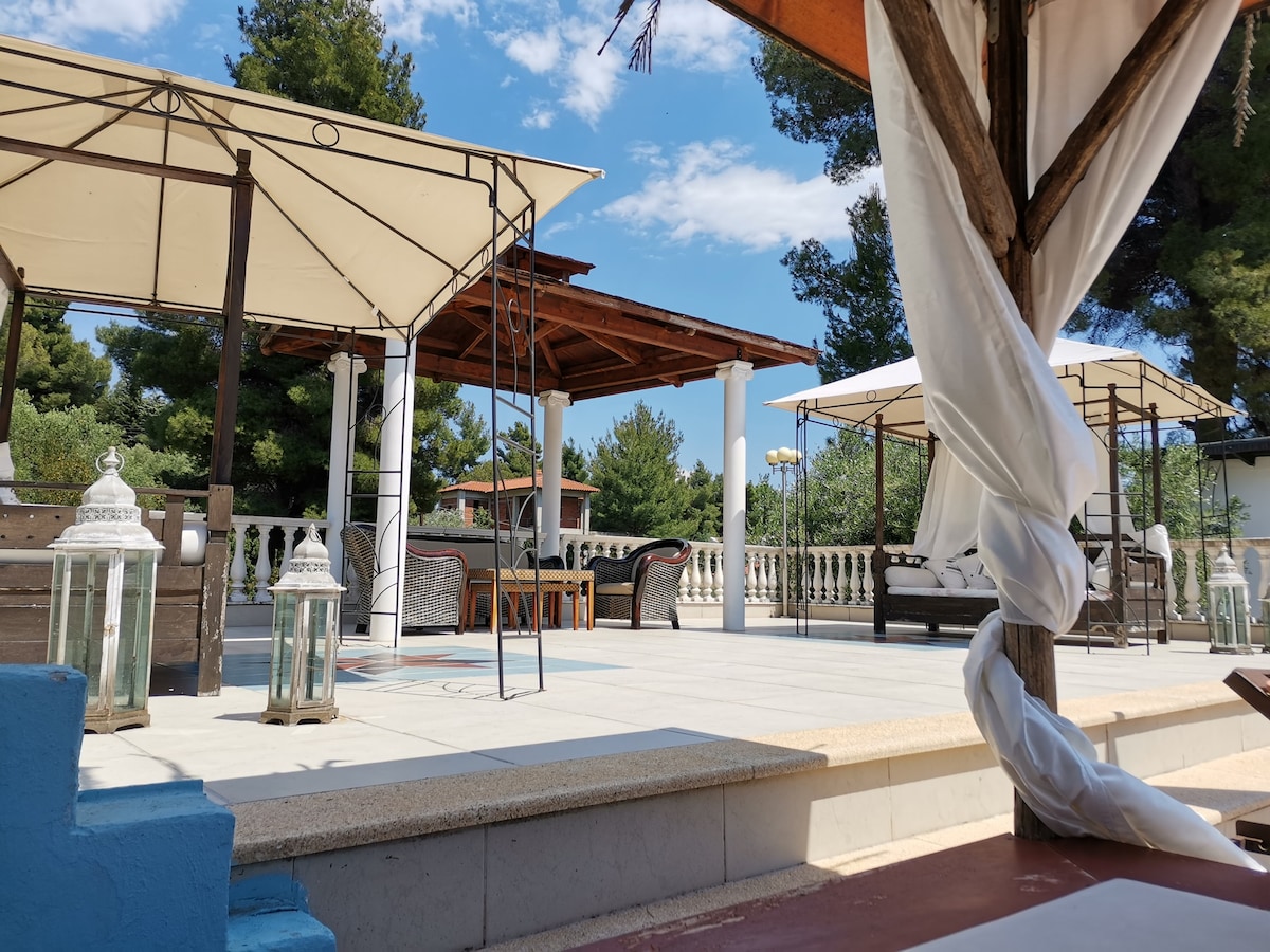 A spacious outdoor area features covered seating arrangements beneath large canopies. Stylish lanterns are placed on the ground, and a lightly decorated railing can be seen in the background. A clear blue sky and greenery enhance the tranquil setting.
