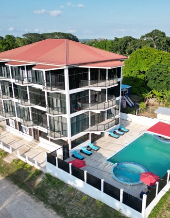 An aerial view of a modern four-story building showcases a pristine pool surrounded by lounge chairs. The structure features large glass windows and a red roof, with greenery visible in the background, creating a serene atmosphere.