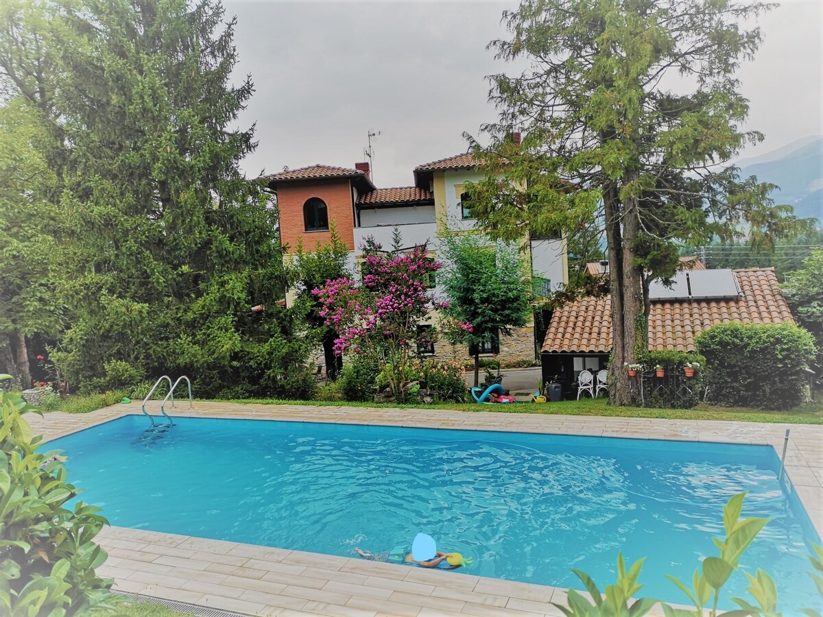 An outdoor swimming pool is surrounded by lush greenery and flowering plants. The clear water of the pool reflects the surrounding trees and buildings, with a patio area visible nearby. The two-story villa features a tiled roof and is partially shaded by the trees.