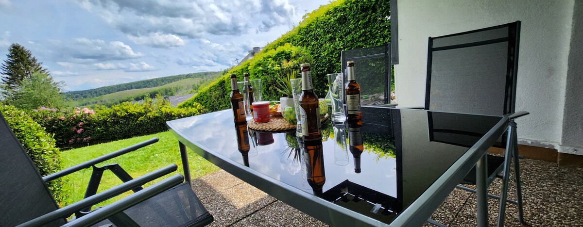 A glass-top table is set outdoors, surrounded by several chairs, with bottles arranged on its surface. The backdrop features a lush green landscape and a vibrant hedge, under a cloudy sky. A serene outdoor setting is created for relaxation.