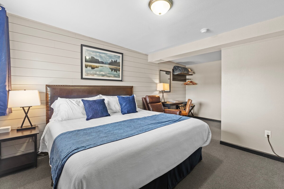 A spacious hotel room features a king-sized bed with a blue accent blanket. Soft natural light enters through a window draped with dark curtains. A bedside lamp and a small desk with a chair are included in the room, providing a functional and welcoming environment.