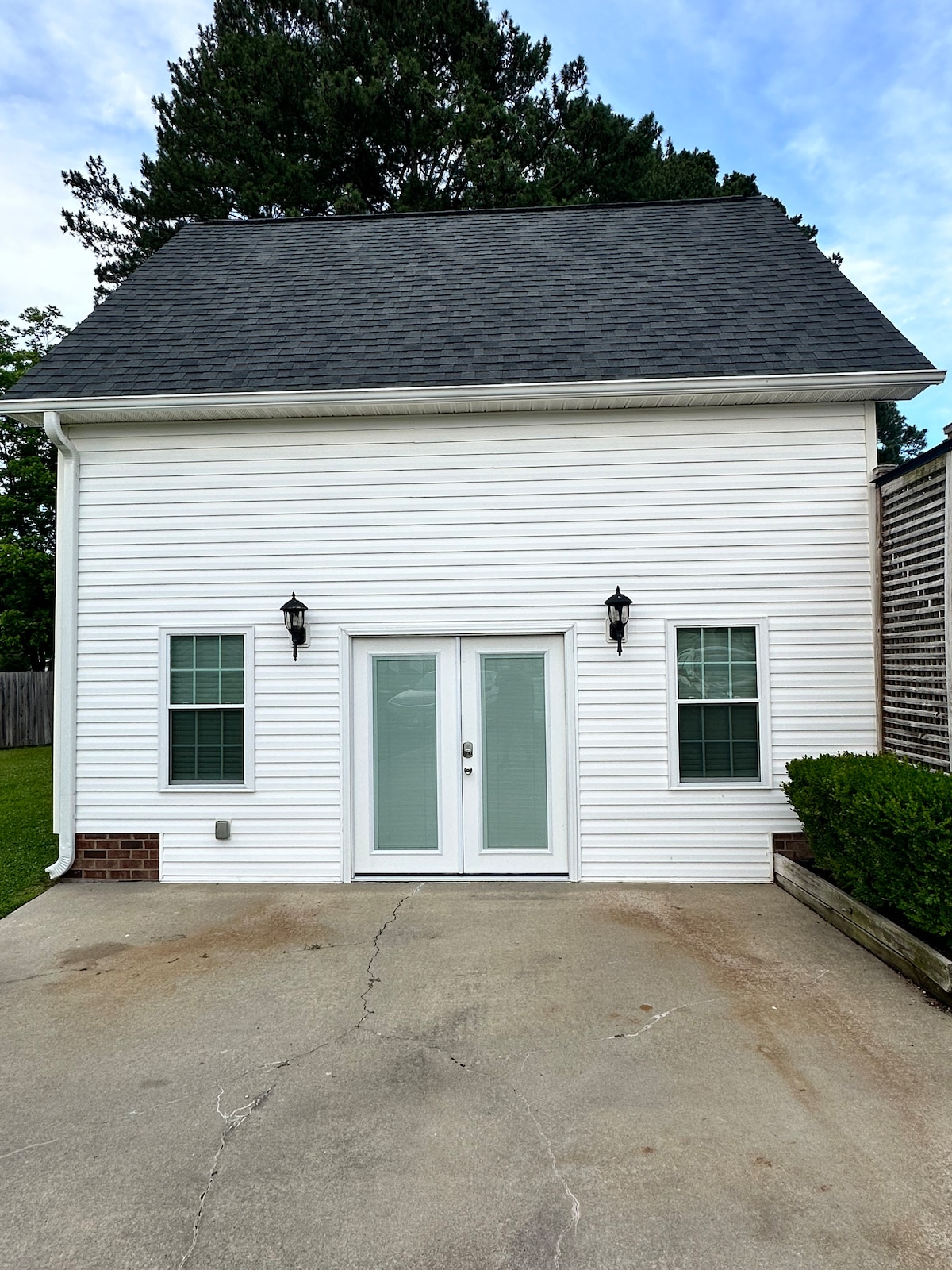 The exterior of the guest house features a clean white siding, flanked by two symmetrical windows on either side of a set of double doors. Lush green landscaping is visible near the entry, under a partly cloudy sky.