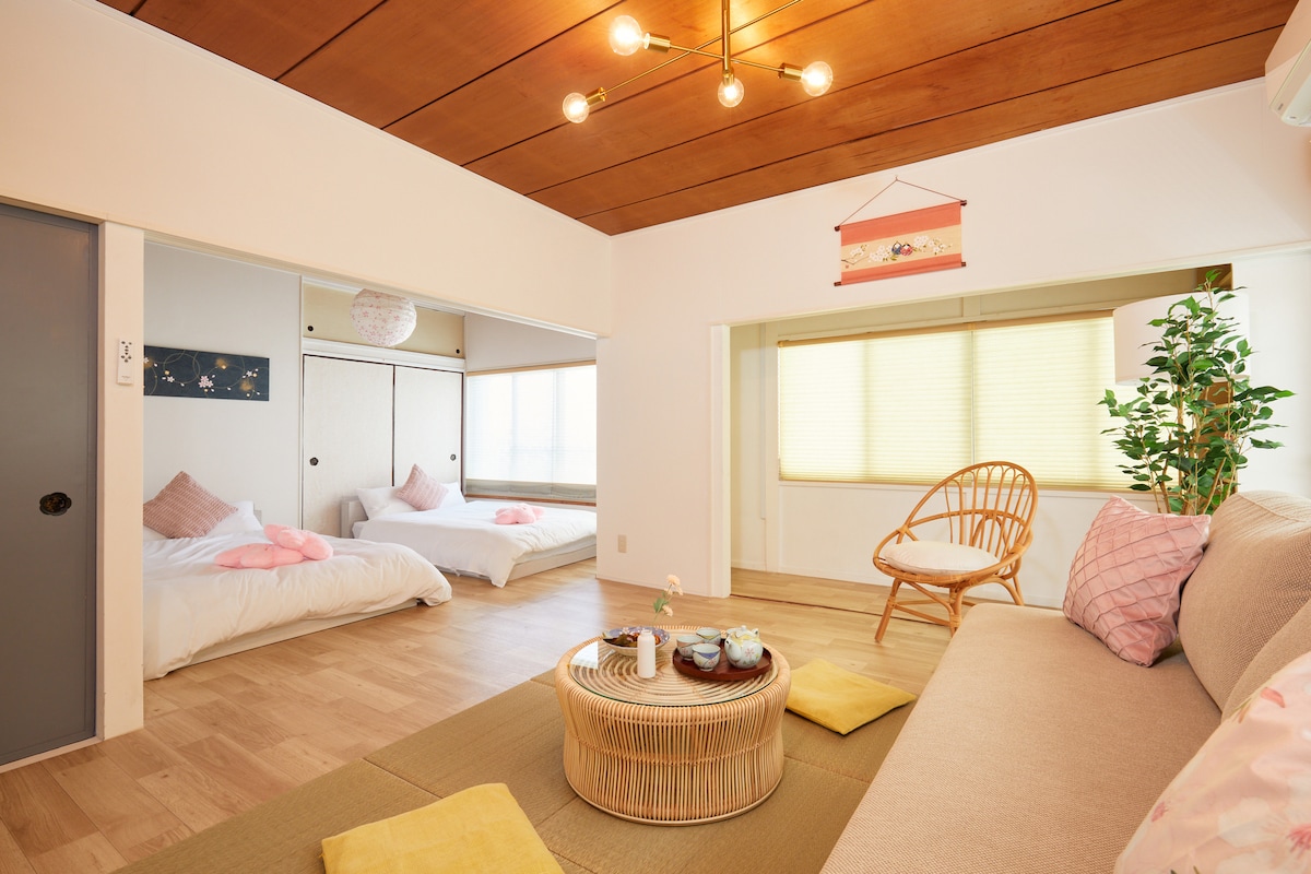 A spacious living area features a comfortable sofa and a round coffee table arranged with tea set. Two bright sleeping areas are visible, furnished with soft bedding. Large windows provide natural light, while decorative elements enhance the inviting ambiance.
