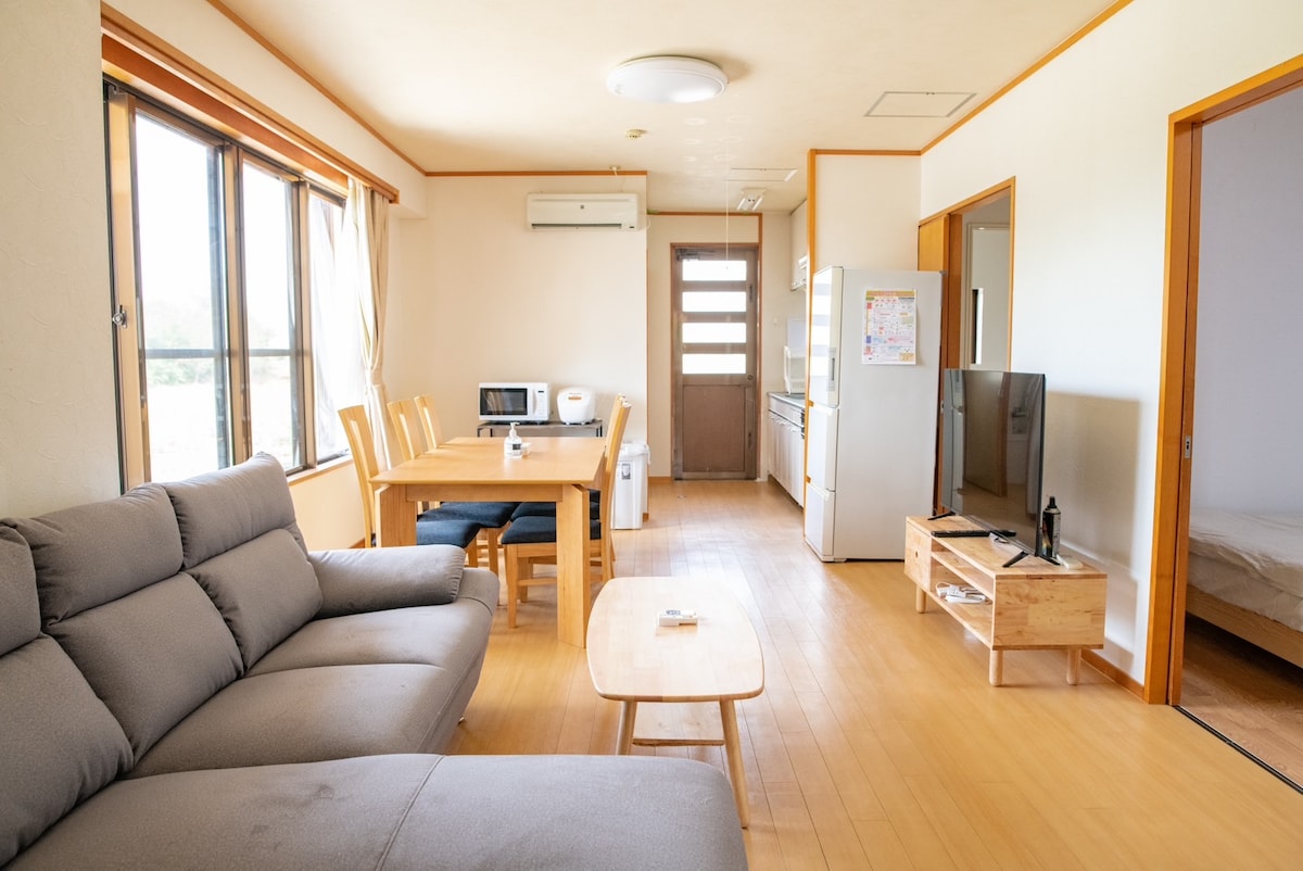 A spacious living area is revealed, featuring a comfortable gray sofa, a wooden coffee table, and a dining table with chairs. Large windows allow natural light to fill the room, providing views of the surrounding landscape. A TV and kitchen appliances are also visible.