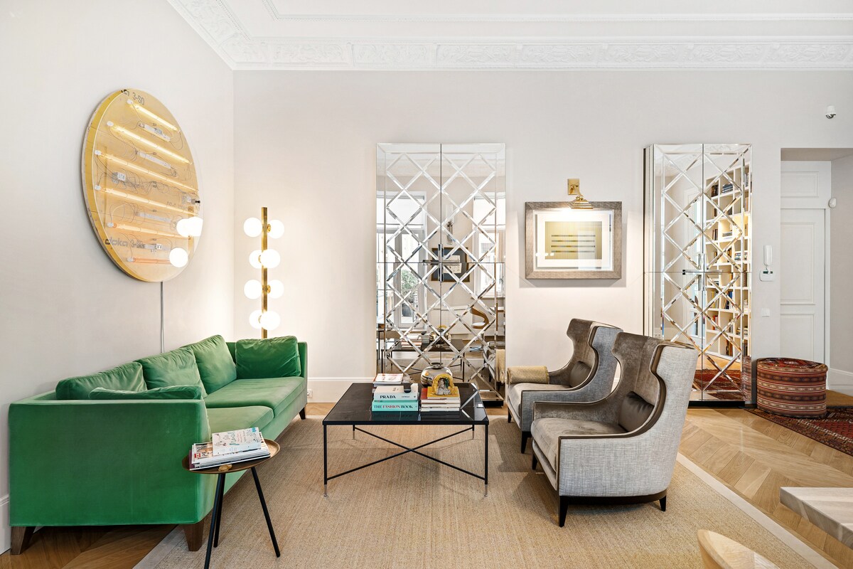 A stylish living area features a green sofa and two gray armchairs arranged around a black coffee table. Coordinated lighting fixtures provide soft illumination, while two decorative mirrors reflect the open space, enhancing the modern design.
