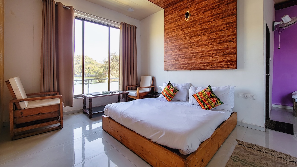 The room features a spacious bed with crisp white bedding, complemented by colorful decorative cushions. Two wooden chairs are positioned beside a small table. A large window allows natural light to fill the space, with views of nature visible through the sheer curtains.