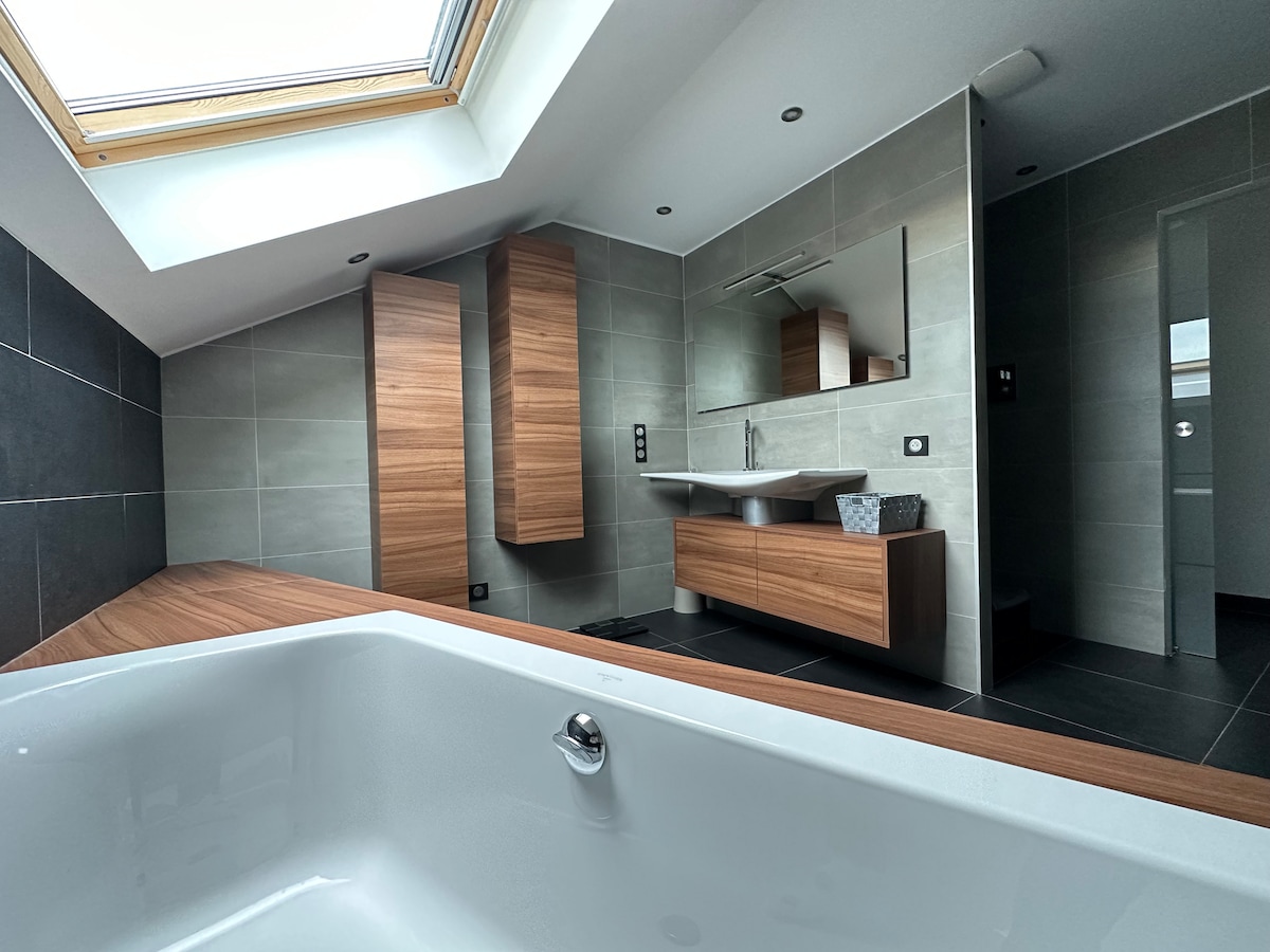 A modern bathroom is showcased with a spacious bathtub positioned in the foreground. Wooden cabinetry complements sleek gray tiles, while a large skylight illuminates the area. A contemporary sink with a wall-mounted mirror is visible, enhancing the overall minimalist design.