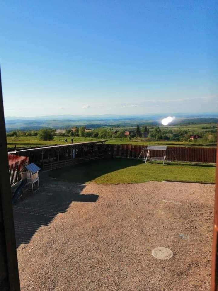 A view of rolling hills and distant mountains is captured from an open space. The yard features a grassy area and a swing set, with a dirt pathway leading to a shaded seating area. The clear blue sky enhances the serene landscape.