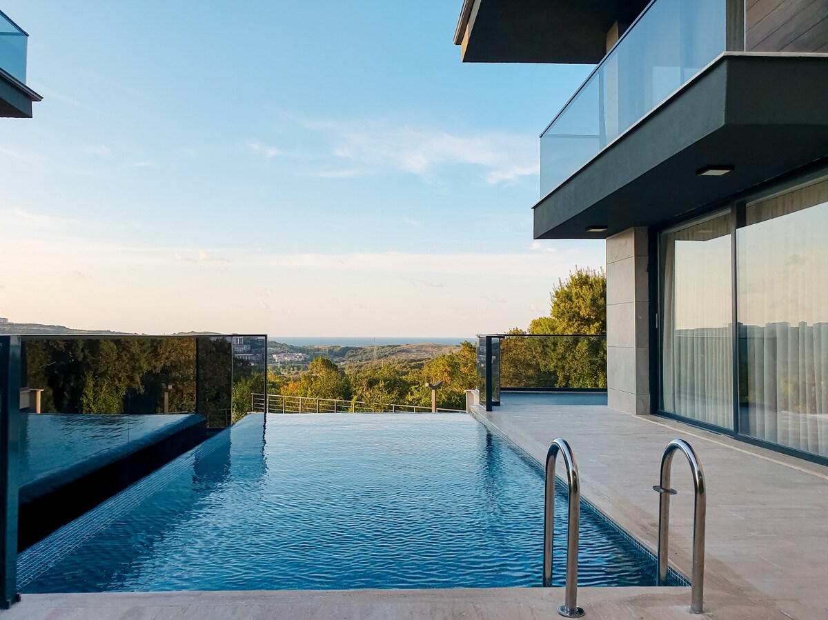 A modern outdoor space is featured with a crystal-clear pool that reflects the surrounding greenery. Glass railings provide an unobstructed view of the landscape, while sleek architecture enhances the serene atmosphere. The area is illuminated by natural light from the clear blue sky.