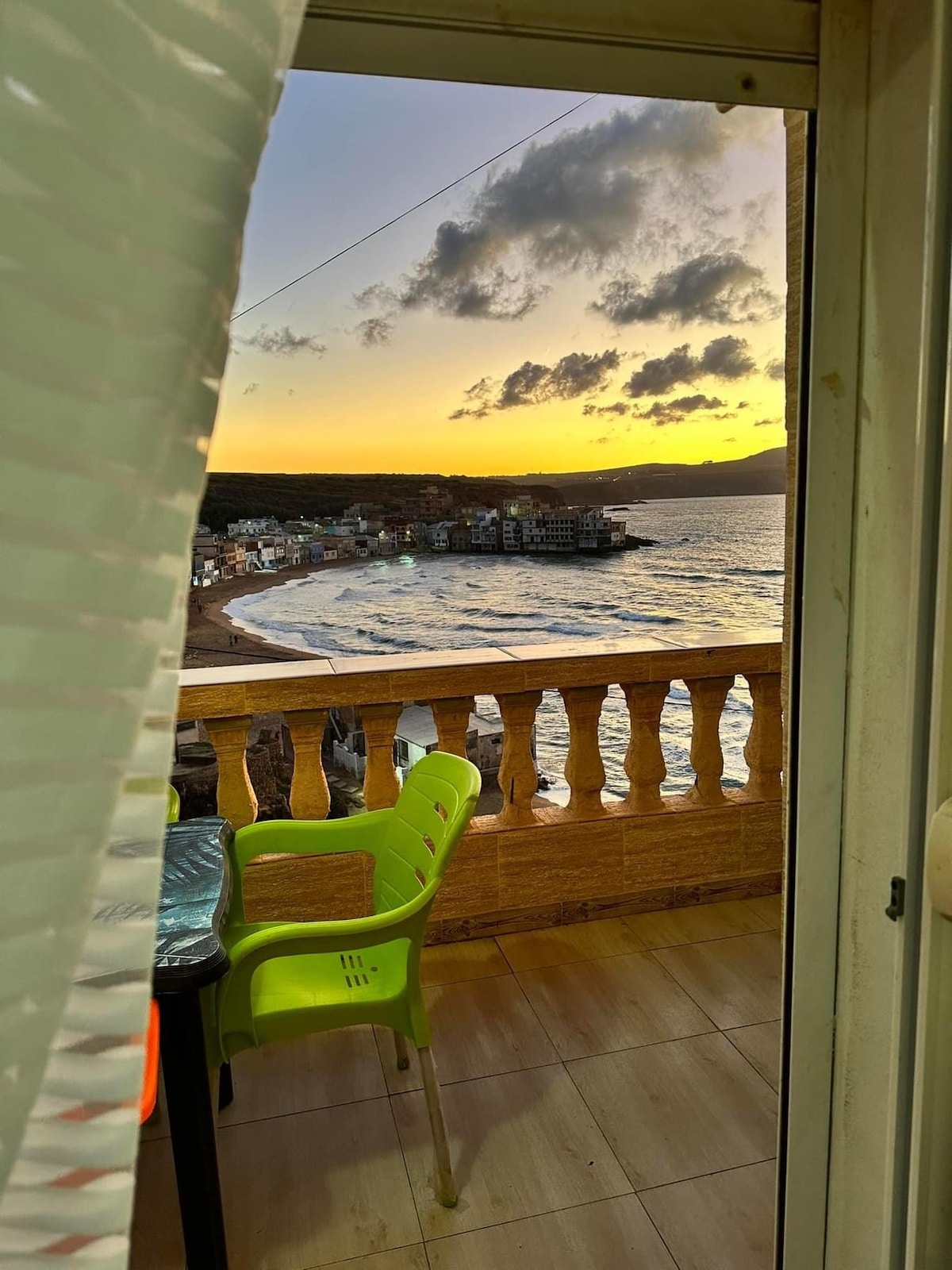 A view from a balcony showcases a vibrant sunset over the beach. Waves gently lap against the shore, while colorful buildings line the coastline. Green outdoor chairs are visible on the balcony, providing a place to relax and enjoy the scenic landscape.