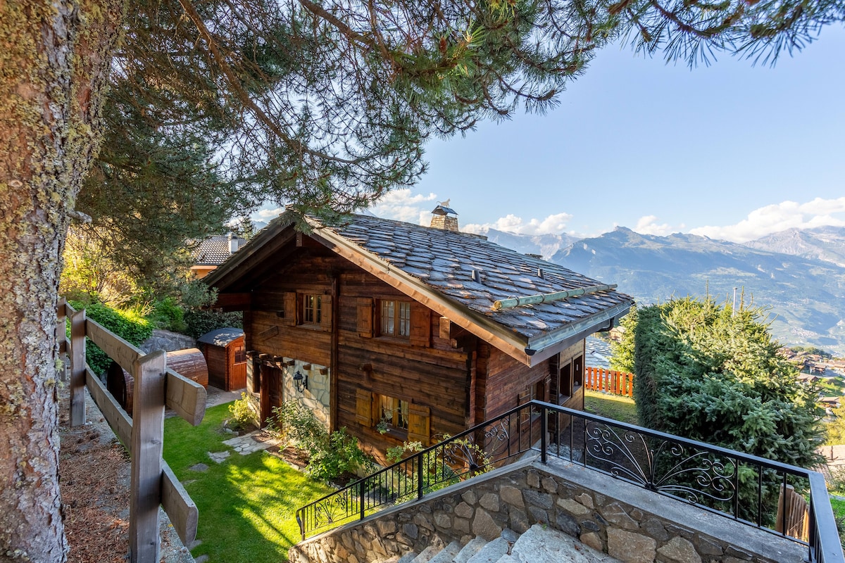 The rustic chalet is framed by lush greenery and features a stone-paved staircase leading to the entrance. A sloped roof with wooden shingles complements the natural surroundings, while expansive windows offer views of the mountainous landscape. A small terrace is visible atop the staircase.
