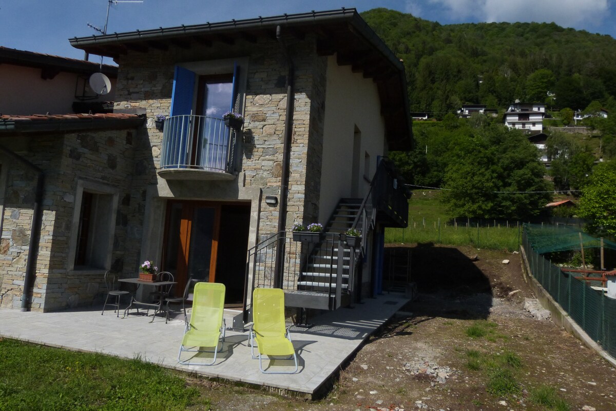 The exterior of the villa is displayed, showcasing a stone façade and a balcony with blue window shutters. A staircase leads to the upper floor, while two bright yellow loungers are positioned on the patio, inviting relaxation amidst the surrounding greenery.