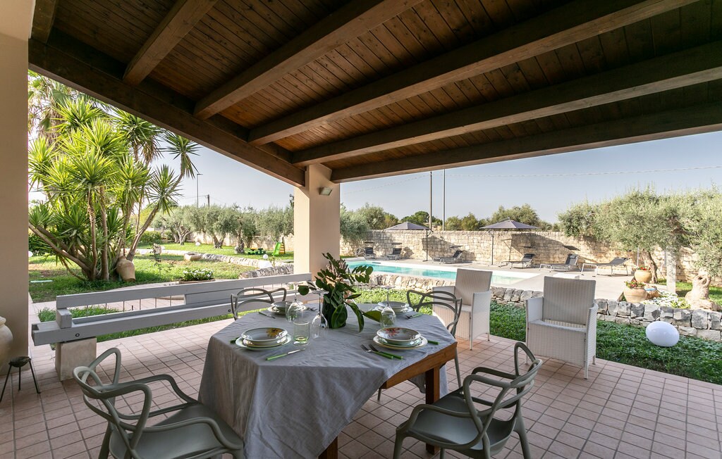 An outdoor dining area features a wooden table set with plates and cutlery. The space is shaded by a wooden ceiling, surrounded by greenery, including palm trees and olive trees. A modern pool is visible in the background, enhancing the tranquil setting.