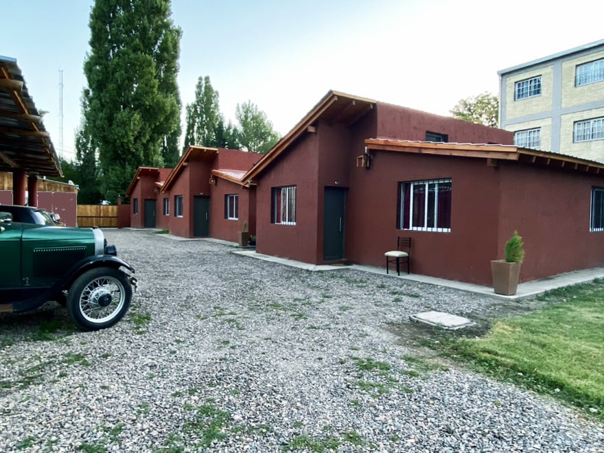 Three rustic buildings with peaked roofs are arranged along a gravel pathway. Each structure features large windows with black frames. A classic car is parked in the foreground, alongside potted plants. Lush greenery surrounds the area, creating a serene outdoor setting.