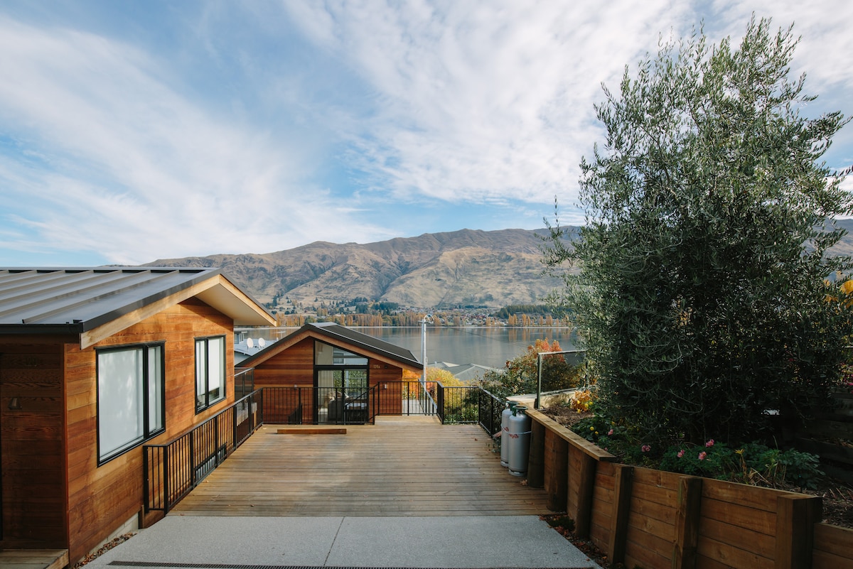 An outdoor view showcases the wooden exterior of the chalet with expansive deck space. The area is surrounded by greenery and features the tranquil Lake Wanaka in the background, framed by mountains and a blue sky with scattered clouds.