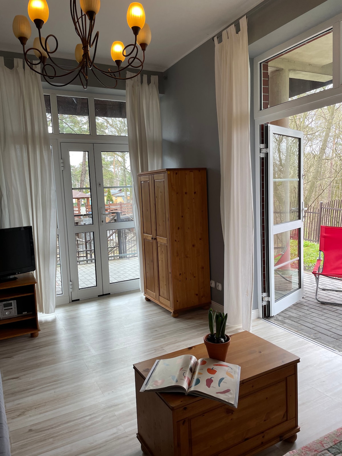 A bright living area features a wooden coffee table and a sleek cabinet. Large windows with sheer curtains provide natural light and views of the outdoors. A television is positioned on a shelf, while a small potted plant adds a touch of greenery.