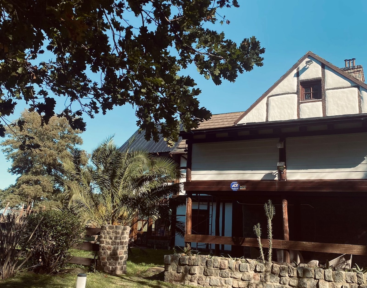 A charming house with a wooden porch is surrounded by lush greenery, including a palm tree. Natural stone elements are visible in the landscape, adding texture. The exterior features a combination of white and wooden accents, set against a bright blue sky.