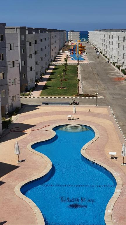 A long, winding pool is featured at the center of a spacious courtyard, surrounded by palm trees and lounge chairs. Adjacent areas include colorful recreational structures and well-maintained pathways leading toward the ocean in the distance.