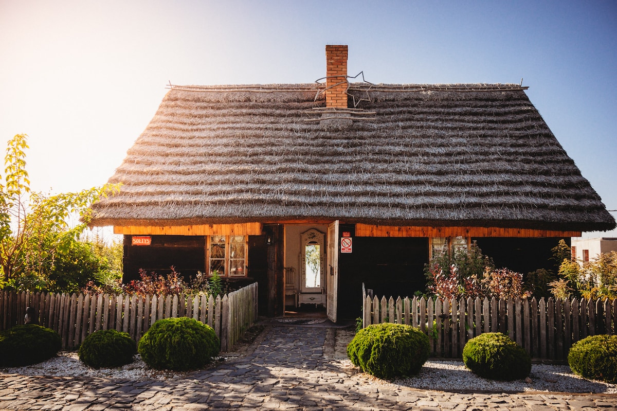 A charming thatched-roof cottage is framed by a wooden fence and landscaped gardens. The exterior features dark wooden panels and a welcoming entrance. Flowering shrubs and neatly trimmed bushes create a cozy outdoor ambiance, complemented by the sun casting a warm glow on the structure.