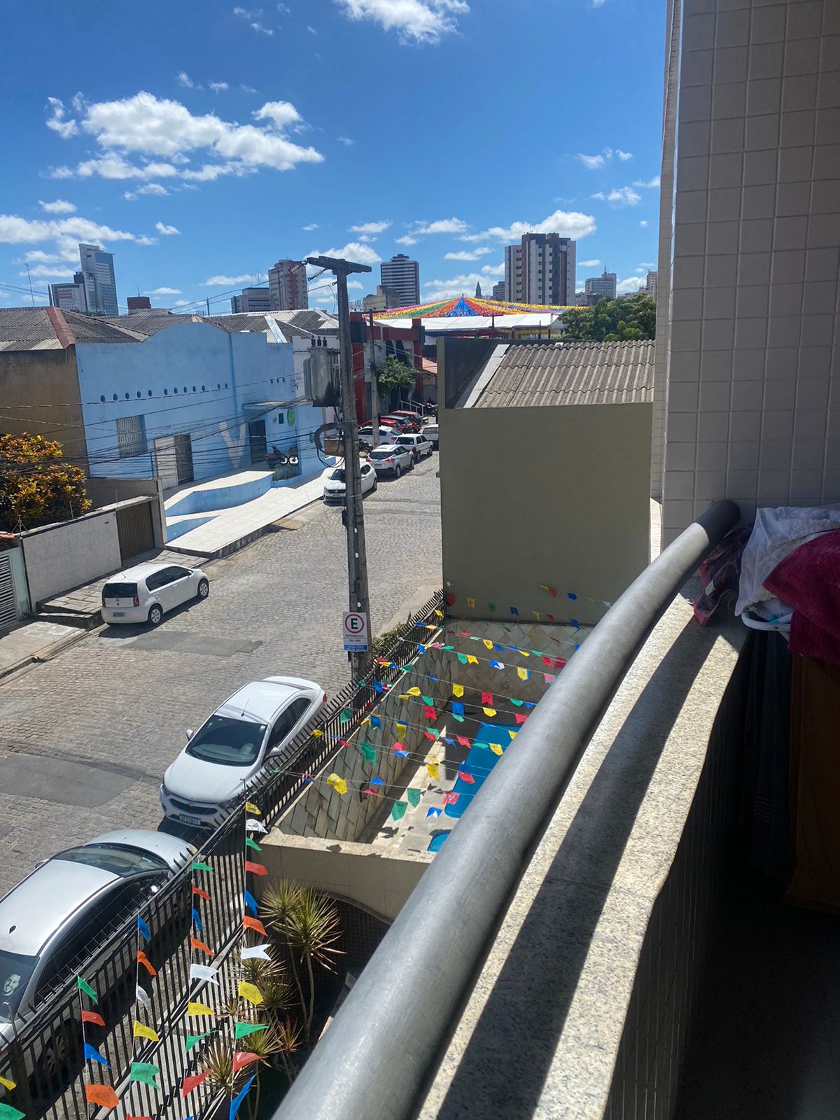 A balcony view showcases a lively street below, where colorful flags are displayed along the railing. The scene is complemented by nearby buildings and a clear blue sky dotted with a few clouds. Urban life is visible with parked cars and the distant backdrop of city structures.