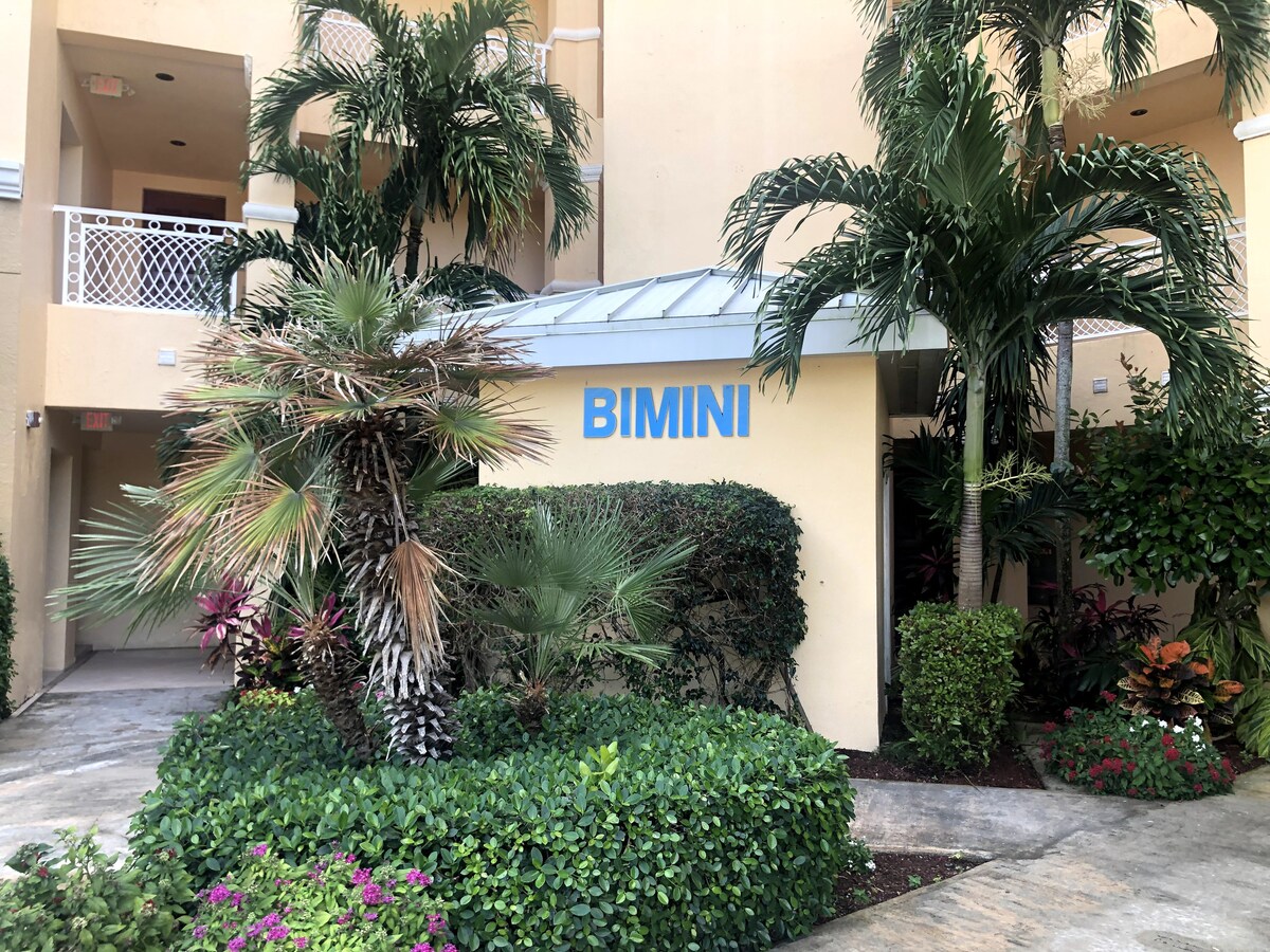 The entrance to the Bimini complex is framed by lush tropical landscaping, featuring a variety of palms and vibrant foliage. A sheltered canopy provides protection from the elements, and the welcoming sign displays the name 'BIMINI' prominently.
