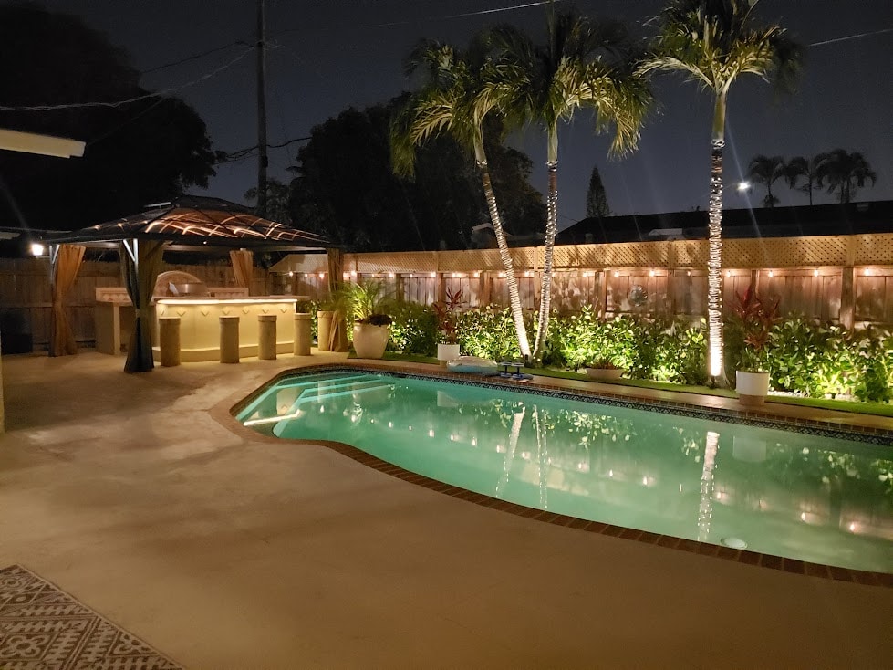 A serene pool area features soft lighting illuminating lush landscaping and palm trees. A covered outdoor bar with seating complements the inviting turquoise waters of the pool, creating a relaxed evening atmosphere.