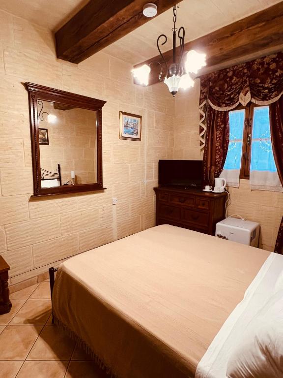 A cozy double room is presented with a warm-toned bedspread covering a spacious bed. A wooden dresser and flat-screen TV are visible, complemented by a mirror reflecting the room's ambiance. Natural light filters through the window, adorned with patterned curtains, enhancing the inviting atmosphere.