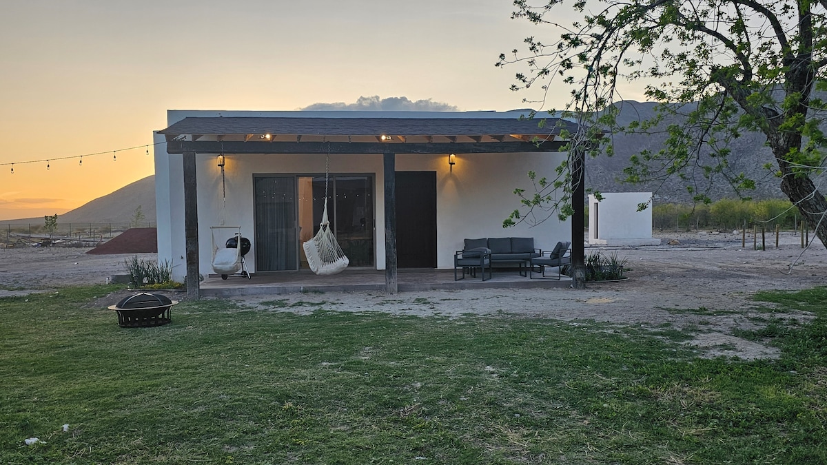 A peaceful exterior view of the villa is presented, featuring a covered porch with comfortable seating and a hanging chair. The grassy area is positioned in front, with soft lighting suspended overhead and mountains visible in the background, creating a relaxed atmosphere for outdoor gatherings.