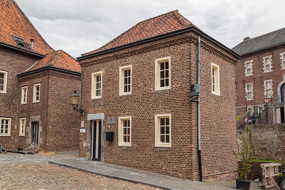 A charming brick building features a classic design with a gabled roof and white-framed windows. The structure is situated on a cobblestone path, surrounded by traditional architecture, and offers a sense of historic elegance.