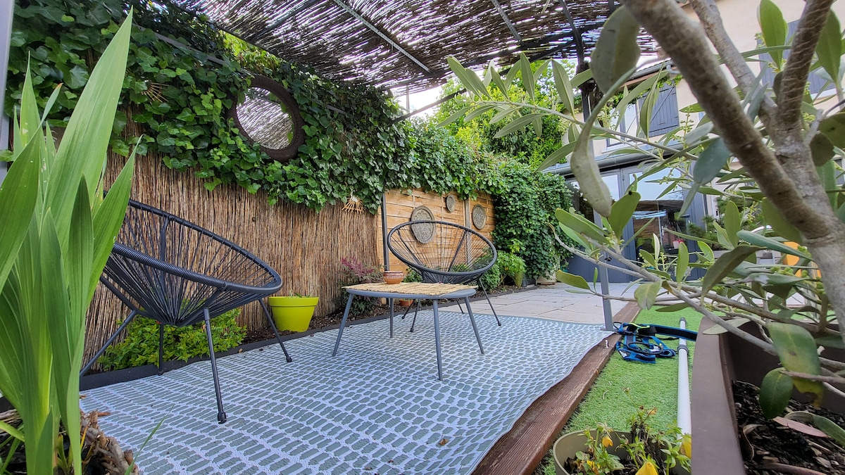 An inviting outdoor space features two modern chairs and a low table set on a patterned rug. Lush greenery surrounds the area, complemented by a decorative wooden wall and potted plants. A shaded canopy provides a comfortable retreat from the sun.