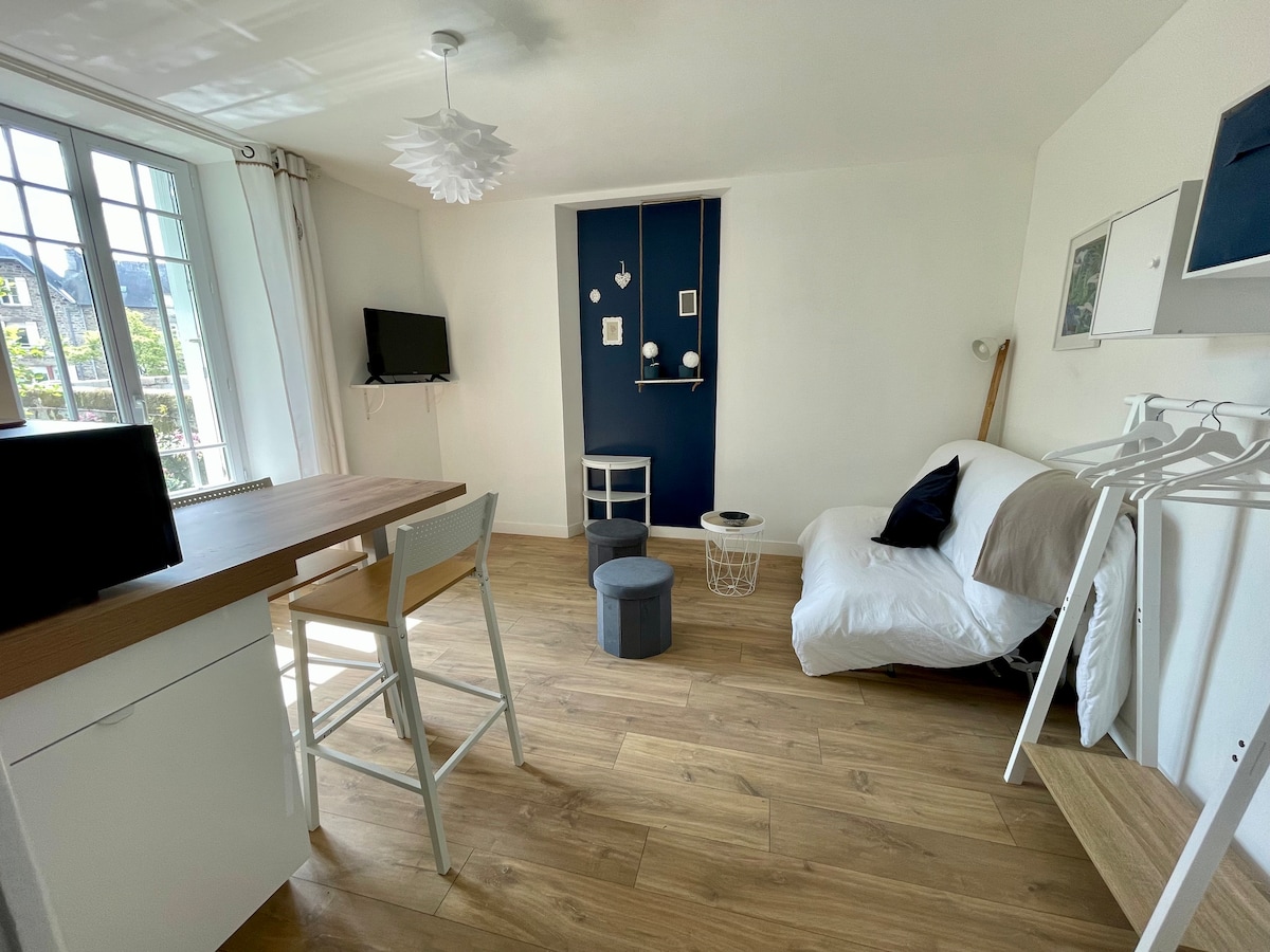 A compact studio is shown with light wood flooring and soft white walls. A small kitchenette is positioned to one side, while a seating area features a white sofa and an accent chair. Natural light enters through large windows, enhancing the bright and airy ambiance.