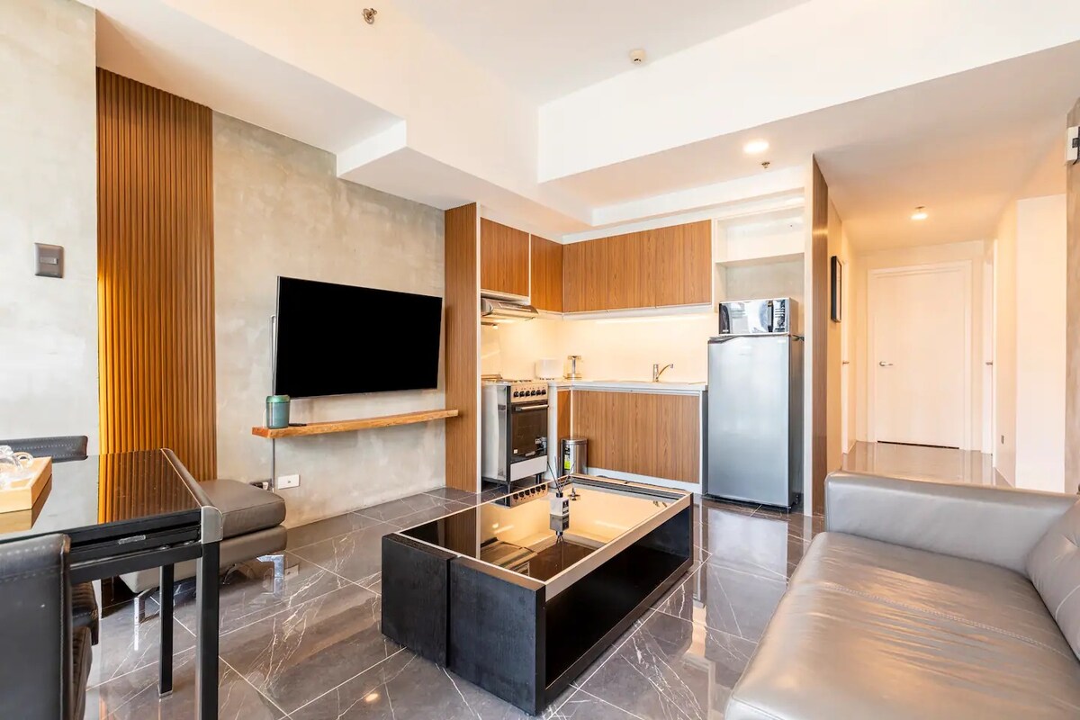 The open-concept living area features a sleek, modern design with a comfortable gray sofa. A glass coffee table sits centrally on a marble floor. The kitchen area is equipped with wooden cabinetry, a stove, and modern appliances, all illuminated by soft lighting.