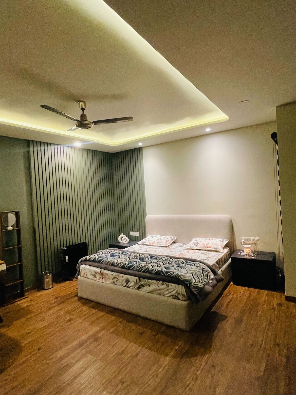 A well-lit bedroom features a spacious bed with a patterned blanket and matching pillows. Soft lighting highlights the smooth walls and textured accents. A nightstand is positioned beside the bed, and wooden flooring adds warmth to the space.